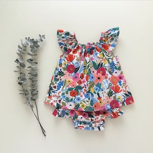 May include: A white baby dress with a floral print in shades of red, pink, blue, and yellow. The dress has a ruffled neckline and a gathered skirt. It is paired with matching bloomers.