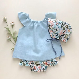 May include: A light blue baby dress with ruffled sleeves and a floral print bonnet. The dress has a gathered neckline and a simple design. The bonnet has a floral print with a variety of colors, including pink, orange, and green.