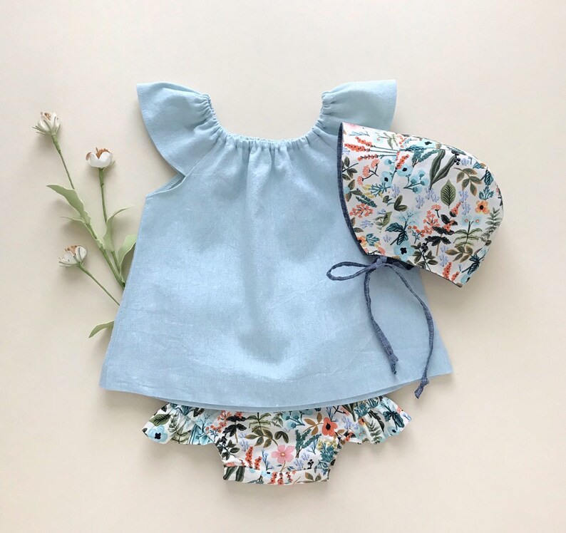 May include: A light blue baby dress with ruffled sleeves and a floral print bonnet. The dress has a gathered neckline and a simple design. The bonnet has a floral print with a variety of colors, including pink, orange, and green.