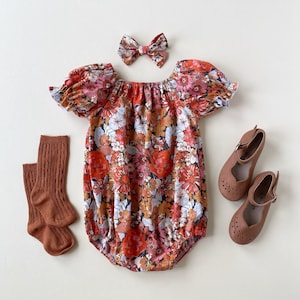 May include: A floral print baby romper with matching bow, brown socks, and brown Mary Jane shoes. The romper features a vibrant pattern of red, orange, pink, and white flowers on a dark background. The shoes have a cut-out design.
