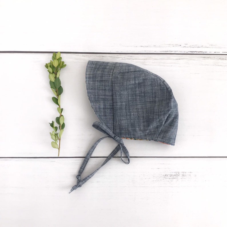 May include: A blue denim baby bonnet with a floral print lining. The bonnet has a tie at the back.