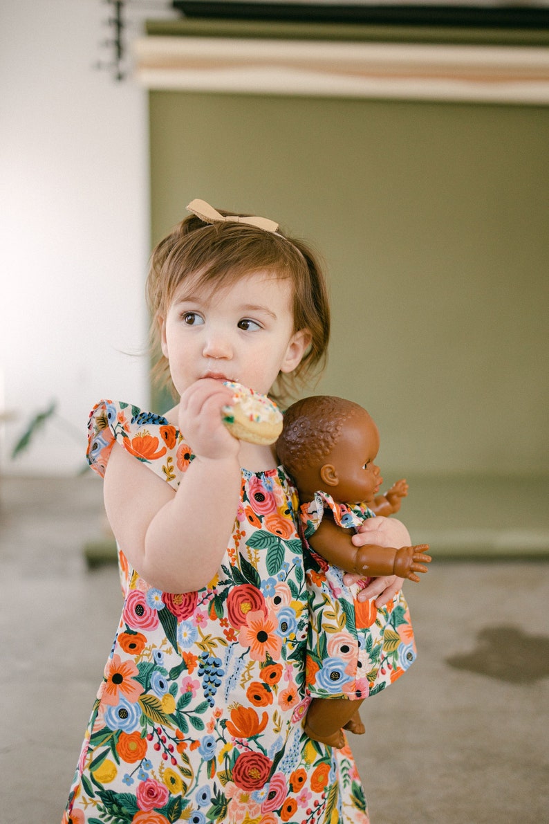 May include: A young child wearing a floral print dress, holding a doll with a matching dress. The dress features a Rifle Paper Co pattern with colorful shades of red, pink, yellow and blue flowers on a cream background. The child is also eating a donut.