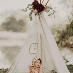 K&ouml;nnte beinhalten: Ein kleines M&auml;dchen in einem Blumenh&uuml;tchen und einem rosa Kleid sitzt in einem wei&szlig;en Tipi, das mit burgunderfarbenen und rosafarbenen Blumen geschm&uuml;ckt ist. Das Tipi ist im Freien auf einem wei&szlig;en, flauschigen Teppich aufgebaut.