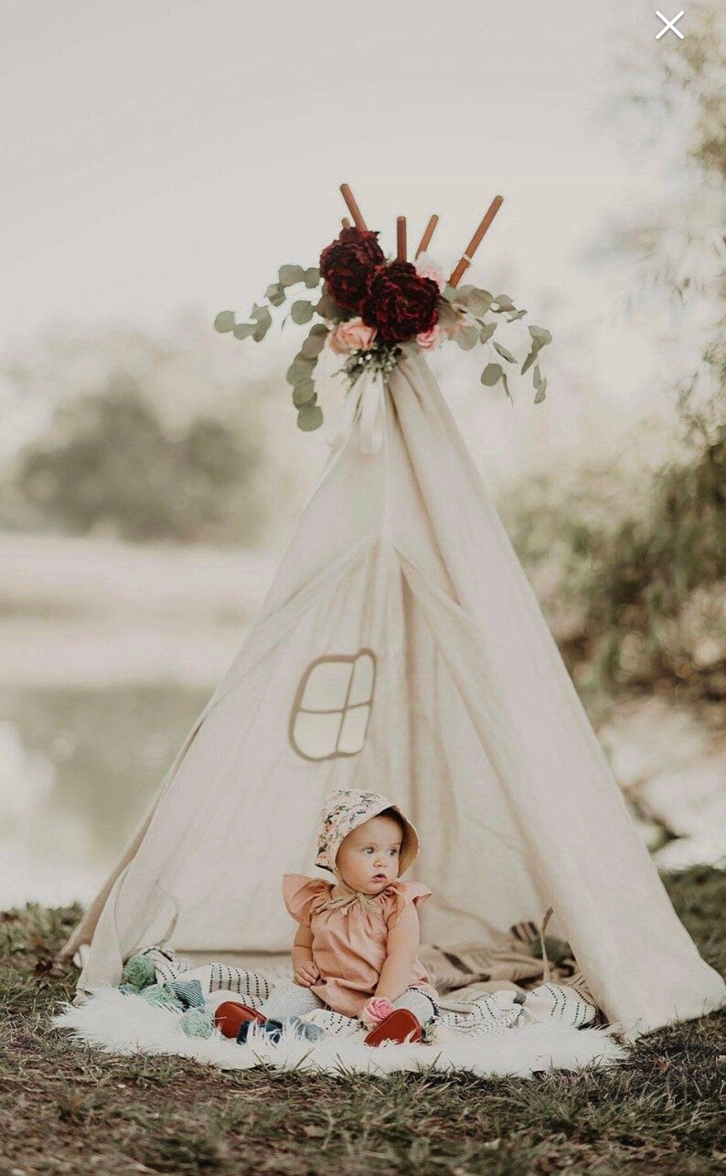 K&ouml;nnte beinhalten: Ein kleines M&auml;dchen in einem Blumenh&uuml;tchen und einem rosa Kleid sitzt in einem wei&szlig;en Tipi, das mit burgunderfarbenen und rosafarbenen Blumen geschm&uuml;ckt ist. Das Tipi ist im Freien auf einem wei&szlig;en, flauschigen Teppich aufgebaut.