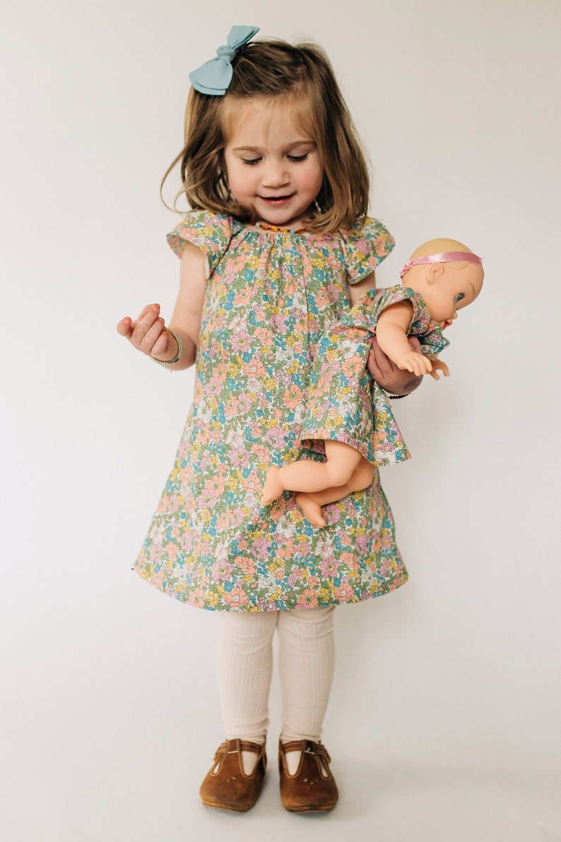 May include: A floral print dress with flutter sleeves and a matching doll dress. The fabric features a pattern of small pink, yellow, and blue flowers on a green background. The dress has a round neckline and a loose, A-line silhouette.