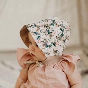 K&ouml;nnte beinhalten: Ein Baby tr&auml;gt einen wei&szlig;en Sonnenschutzhut mit Blumenmuster. Der Hut hat einen braunen Rand und gr&uuml;ne und rosa Blumen. Das Baby tr&auml;gt ein rosa Kleid mit R&uuml;schen&auml;rmeln.
