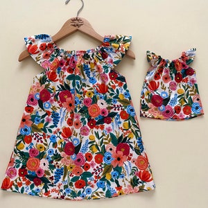 May include: A matching set consisting of a dress and a smaller top, both featuring a white background with a vibrant floral print in shades of red, pink, blue, yellow, and green. The garments have flutter sleeves and a gathered neckline.