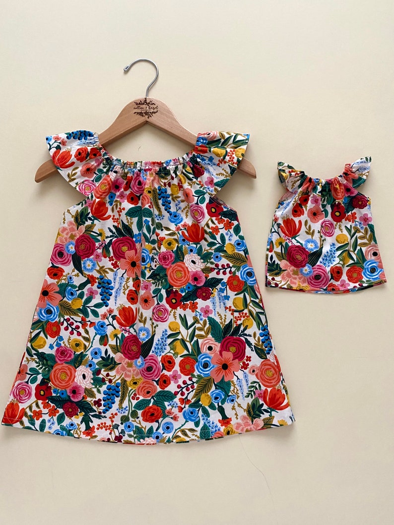 May include: A matching set consisting of a dress and a smaller top, both featuring a white background with a vibrant floral print in shades of red, pink, blue, yellow, and green. The garments have flutter sleeves and a gathered neckline.