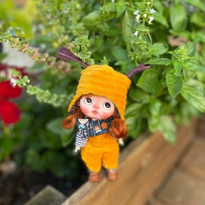May include: A small doll wearing a yellow corduroy hat with brown antlers, a yellow corduroy overall, and a black and white patterned scarf. The doll has red hair and brown eyes.