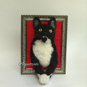 May include: A black and white cat with yellow eyes is peeking out of a silver picture frame with a red background. The cat is made of felt and is a 3D sculpture.