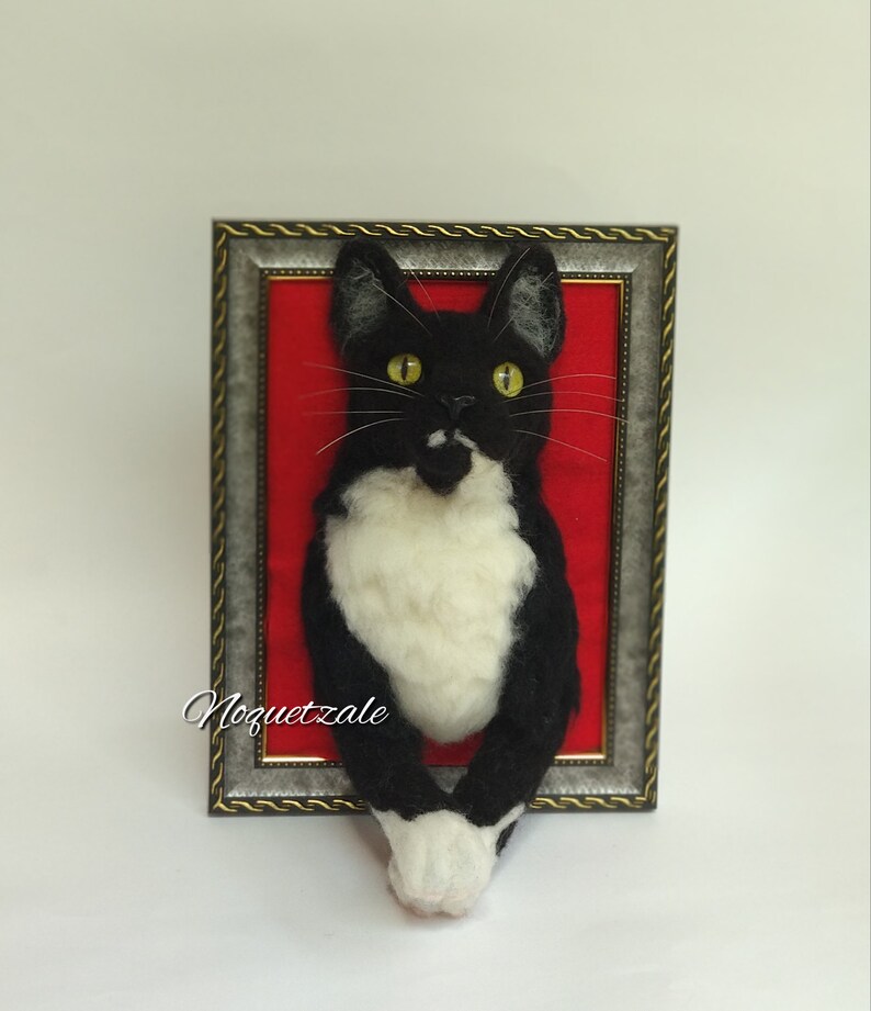 May include: A black and white cat with yellow eyes is peeking out of a silver picture frame with a red background. The cat is made of felt and is a 3D sculpture.