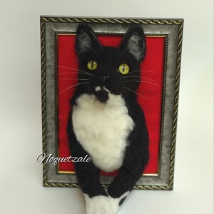 May include: A black and white cat with yellow eyes is depicted in a framed needle felted artwork. The cat is posed with its front paws sticking out of the frame. The background is a red felt.