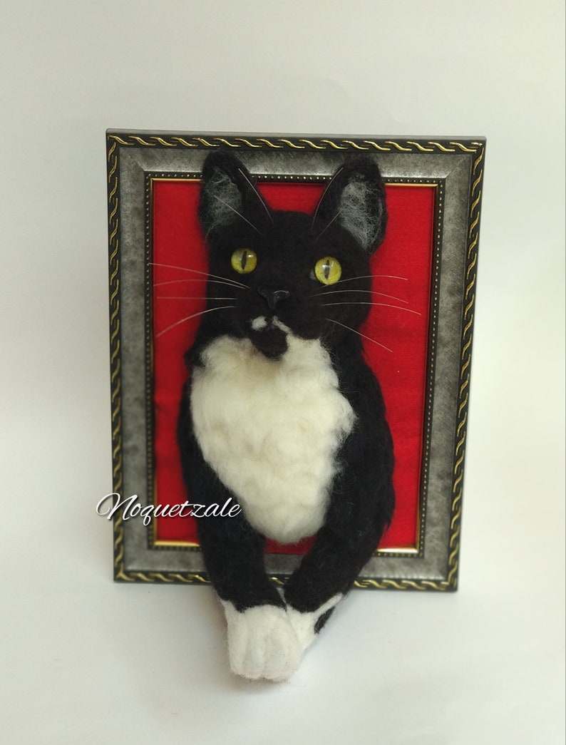 May include: A black and white cat with yellow eyes is depicted in a framed needle felted artwork. The cat is posed with its front paws sticking out of the frame. The background is a red felt.