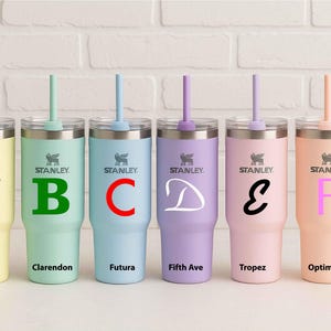 May include: Six Stanley tumblers in pastel colors, each with a different letter and font style: A (Brush), B (Clarendon), C (Futura), D (Fifth Ave), E (Tropez), and F (Optima). Each tumbler has a straw and handle.