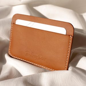 May include: A tan leather card holder with a white card partially visible in the top slot. The card holder has a rounded top and is stitched with white thread. The text "The Gray Deer" is embossed on the front.