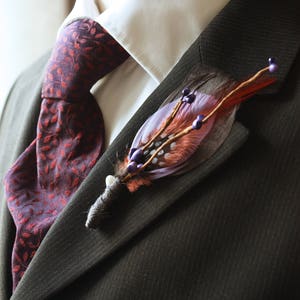 May include: A brown suit jacket with a white shirt and a burgundy patterned tie. A feather and berry boutonniere is pinned to the lapel.