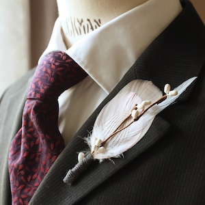 May include: A dark grey suit jacket with a white collared shirt and a burgundy tie featuring a leaf design. A white feather boutonniere with small white details is pinned to the lapel, offering a refined appearance.