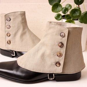 May include: A pair of beige linen gaiters with button closures. The gaiters are worn over black leather shoes with a pointed toe.