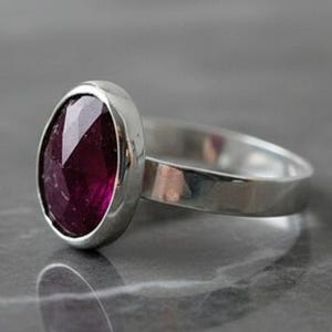 May include: A silver ring with a large, oval-cut, deep red gemstone. The gemstone is set in a bezel setting, and the band is a simple, polished silver. The ring is displayed on a gray, marble-like surface, reflecting the ring's shape.