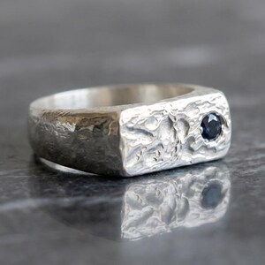 May include: A silver ring with a textured surface and a dark blue gemstone. The ring has a rectangular face with a hammered appearance. The gemstone is set into the face of the ring.