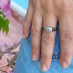 Puede incluir: Un anillo de plata con un diseño de incrustaciones azul y rojo. El anillo se lleva en el dedo índice de una mano.