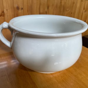May include: A white ceramic chamber pot with a rounded body and a handle. The handle is attached to the side of the pot and has a decorative design. The pot is sitting on a wooden surface.
