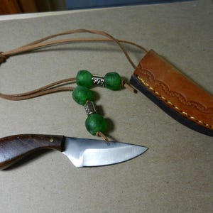 May include: A small, handcrafted knife with a wooden handle and a stainless steel blade. It includes a leather sheath and a beaded lanyard with green beads. The sheath is brown with decorative stitching.
