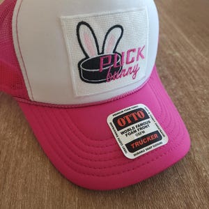 May include: A bright pink and white trucker hat featuring a rectangular patch with an embroidered black hockey puck and white rabbit ears. The text on the patch reads Puck Bunny in pink. A sticker on the brim displays Otto brand information.