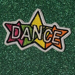 May include: Embroidered patch with the word "DANCE" in white, outlined in black, set against a multi-colored starburst design. The starburst features pink, green, yellow, and orange segments, all bordered by a silver outline. The patch is on a sparkly green background.