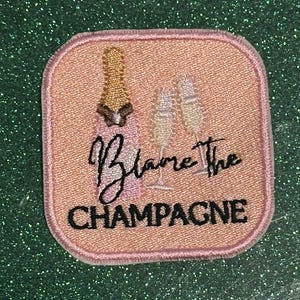 May include: A square, embroidered patch with a pink background features a champagne bottle, two champagne flutes, and the words "Blame the CHAMPAGNE" in black script. The patch has a pink border.