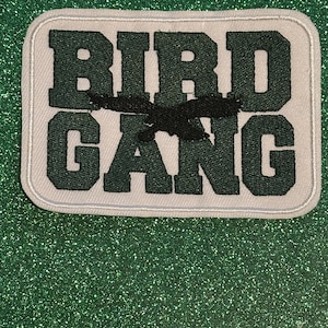 May include: A rectangular patch with the words "BIRD GANG" in dark green block letters. A black bird silhouette is embroidered across the center. The patch has a white background with a stitched border.