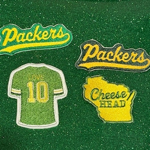 May include: Four embroidered patches featuring Green Bay Packers themes. Two patches display the word Packers in yellow script, one is a green jersey with the name LOVE and number 10, and one is a yellow map of Wisconsin with Cheese Head text.