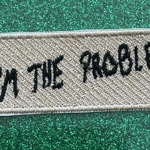 May include: A rectangular patch with the embroidered text "I'M THE PROBLEM" in black on a tan background. The patch has a stitched border and is set against a sparkly green surface.
