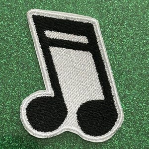 May include: Embroidered patch in the shape of a musical eighth note. The note is black and white with a white border. The patch is on a green glitter background.
