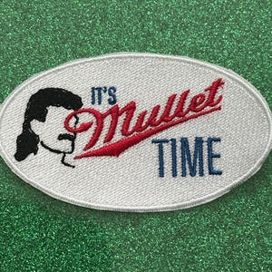May include: Oval-shaped embroidered patch with the text "It's Mullet Time." The patch features a black silhouette of a person with a mullet hairstyle and mustache. The text "Mullet" is in red script, and "TIME" is in blue.