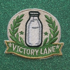 May include: Embroidered patch featuring a milk bottle design, surrounded by green laurel leaves and two small stars. The patch has a tan background and a green banner with the words "VICTORY LANE" in white.