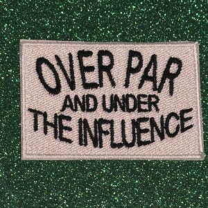May include: A rectangular patch with the text "OVER PAR AND UNDER THE INFLUENCE" embroidered in black on a light gray background. The patch is set against a sparkly, dark green surface.