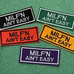 May include: Five rectangular patches in black, green, orange, yellow, and purple, each with the text "MILF'N AIN'T EASY" in white. The patches have a white border and are arranged on a green, glittery surface.