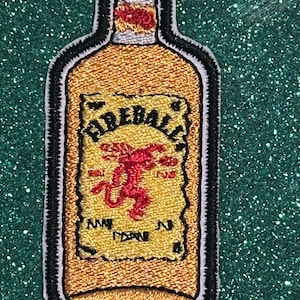 May include: Embroidered patch in the shape of a Fireball cinnamon whisky bottle. The patch features a gold bottle with a red and yellow label, a red dragon, and the word "Fireball". The patch has a black border.