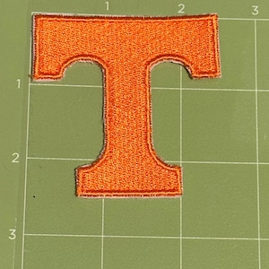 May include: An orange embroidered patch in the shape of the letter "T". The patch has a stitched border and is set against a green grid background. The patch is approximately 2 inches tall and 1.5 inches wide.
