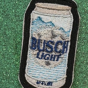 May include: Embroidered patch featuring a can of Busch Light beer. The design includes the brand name in blue lettering, a mountain scene, and a black border. The patch is rectangular with rounded corners.