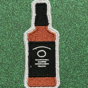 May include: Embroidered patch in the shape of a whiskey bottle. The patch features a brown bottle with a black label and a black top. The label has white embroidered details. The patch is outlined in white.