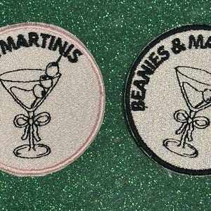 May include: Two round embroidered patches with the text "BEANIE'S & MARTINIS" around a martini glass design. One patch has a pink border, the other a black border. The martini glasses have olives.