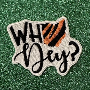 May include: Embroidered patch with the words "WH Dey?" in black script. The patch has a white border and a section with orange and black stripes, resembling a tiger pattern. The patch is on a green glitter background.