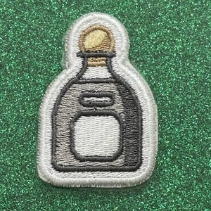 May include: Embroidered patch in the shape of a tequila bottle. The patch features a white border, a gray bottle outline, and a gold-colored cap. The bottle has a rectangular label area.