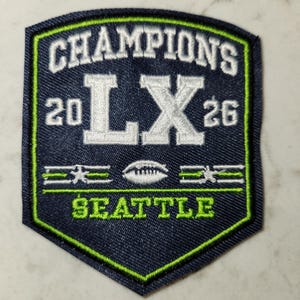 May include: A navy blue embroidered patch with a green and white border. The patch reads "CHAMPIONS LX 26 SEATTLE" in white and green lettering. A football graphic is centered on the patch.