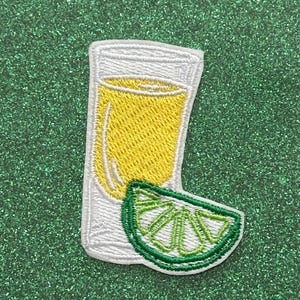 May include: Embroidered patch featuring a shot of tequila with a yellow liquid and a lime wedge. The patch has a white border and is set against a green, glittery background. The tequila shot is in a clear glass.