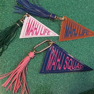 Puede incluir: Cuatro llaveros con forma de banderín con los textos "MAHJ LIFE" y "MAHJ SQUAD". Cada banderín es de un color diferente: blanco, marrón y azul marino. Cada llavero tiene una borla a juego y un cierre dorado.