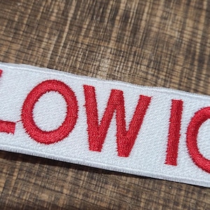 May include: A rectangular white patch with the embroidered text "LOW IQ" in red. The patch is likely designed to be sewn or ironed onto clothing or accessories. The background is a wooden surface.