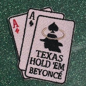 May include: Embroidered patch featuring two playing cards: the Ace of Diamonds and the Ace of Spades. The Ace of Spades card has the text "TEXAS HOLD 'EM BEYONCÉ" in black. The patch is white with black and red details.